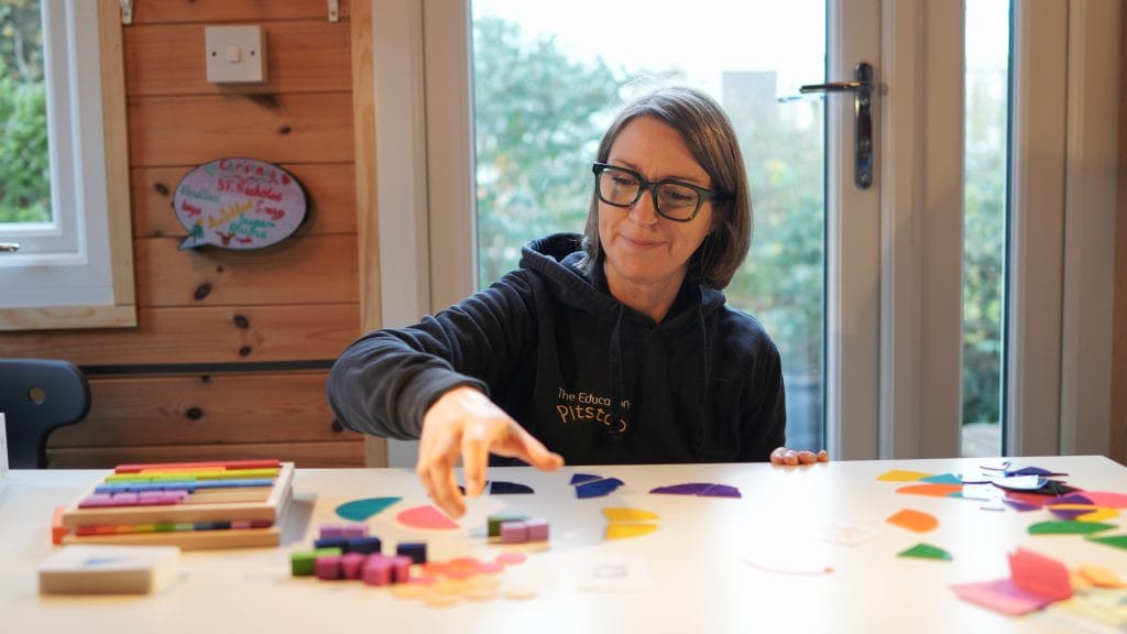 A woman with shoulder-length brown hair and black glasses sits at a white table, reaching toward a variety of colourful educational tools. She is wearing a black hoodie with the text "The Education Pitstop" on the chest. The table is covered with colourful wooden blocks, fraction circles in various colours, and small folded paper slips. Behind her, a wooden wall features a speech-bubble-shaped sign with handwritten names, and a glass door looks out onto a garden.