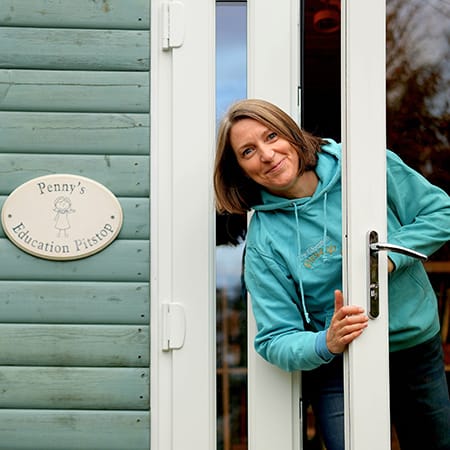 A woman in a teal hoodie smiles and leans out of a white-framed glass door of a light-blue wooden building. To the left of the door is an oval sign that reads "Penny's Education Pitstop" with a small illustration of a child.