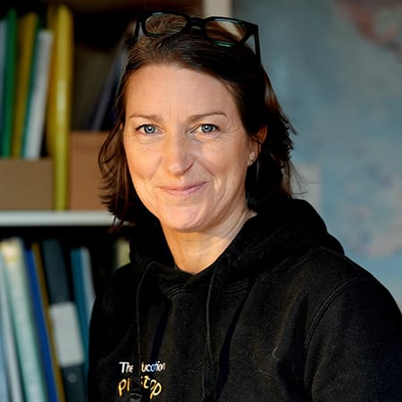A close-up portrait of a Penny Sutherland with shoulder-length brown hair and glasses resting on top of her head. She is wearing a black hoodie that features a small logo on the chest. In the background, a bookshelf filled with colourful binders and a blurred map on the wall are visible.