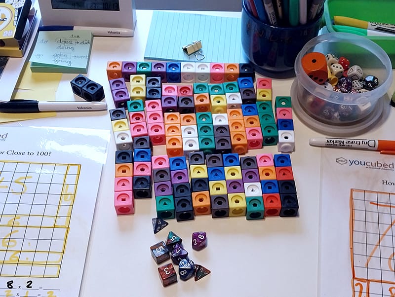 Lessons 1 A high-angle shot of a white desk filled with educational math materials. In the center, there is a large rectangular arrangement of colourful interlocking plastic cubes and several multi-sided polyhedral dice. Two "How Close to 100?" worksheets with hand-drawn grids and numbers in yellow and orange markers are placed on either side. The desk also holds various school supplies, including a blue cup filled with pens and pencils, a green tray with markers, a small digital clock, and a whiteboard with the words "Red = Yellow" written on it.