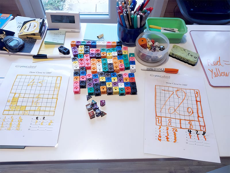 A high-angle shot of a white desk filled with educational math materials. In the center, there is a large rectangular arrangement of colourful interlocking plastic cubes and several multi-sided polyhedral dice. Two "How Close to 100?" worksheets with hand-drawn grids and numbers in yellow and orange markers are placed on either side. The desk also holds various school supplies, including a blue cup filled with pens and pencils, a green tray with markers, a small digital clock, and a whiteboard with the words "Red = Yellow" written on it.