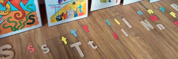 Various wooden and plastic lowercase and uppercase letters are arranged in groups on a light brown wooden floor. The letters include "S," "T," "I," "N," and "P" in different colours and patterns, such as yellow, red, and blue checkered. In the background, several colourful educational posters are leaned against a wall, featuring illustrations like a seahorse and children playing.