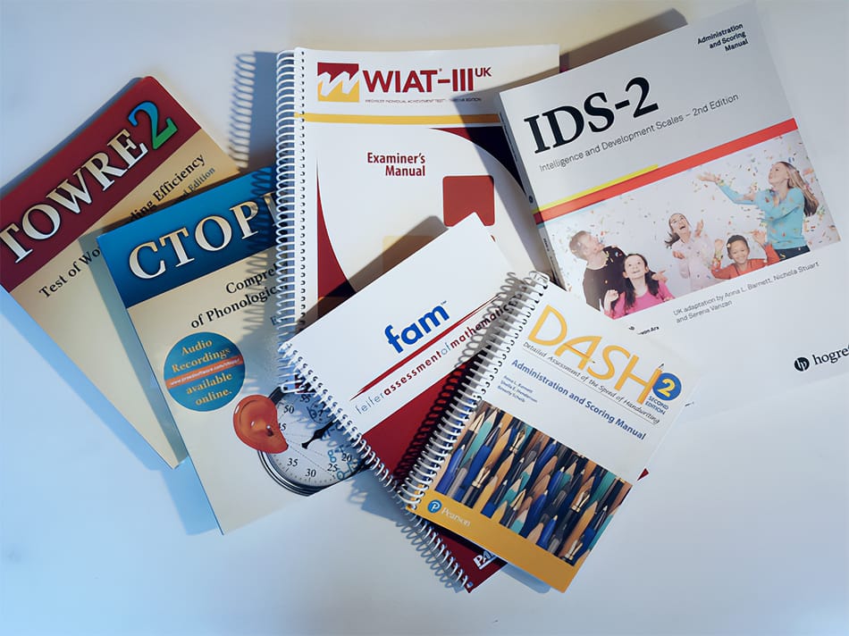 A collection of several educational assessment manuals and spiral-bound books are scattered across a white surface. The visible titles include "TOWRE-2," "CTOPP-2," "WIAT-III UK Examiner's Manual," "fam Feifer Assessment of Mathematics," "DASH-2 Administration and Scoring Manual," and "IDS-2 Intelligence and Development Scales." Several of the books feature logos from publishers like Pearson and Hogrefe.
